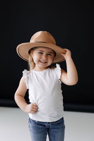A child in a white top and tan hat poses playfully against a black background.