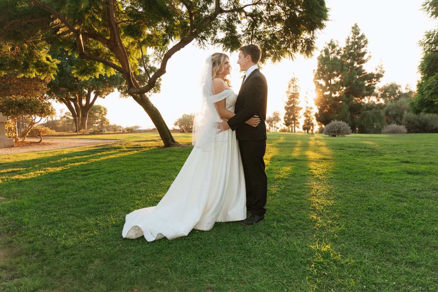 A couple shares a romantic sunset moment on a grassy lawn with golden sunlight filtering through trees.