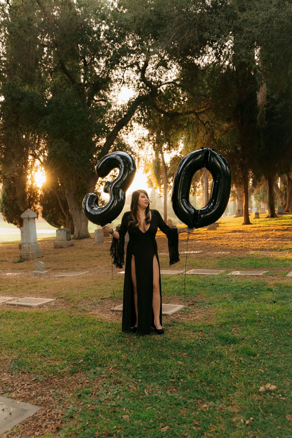 Black-clad figure poses with shiny metallic balloons in a misty park at sunset.