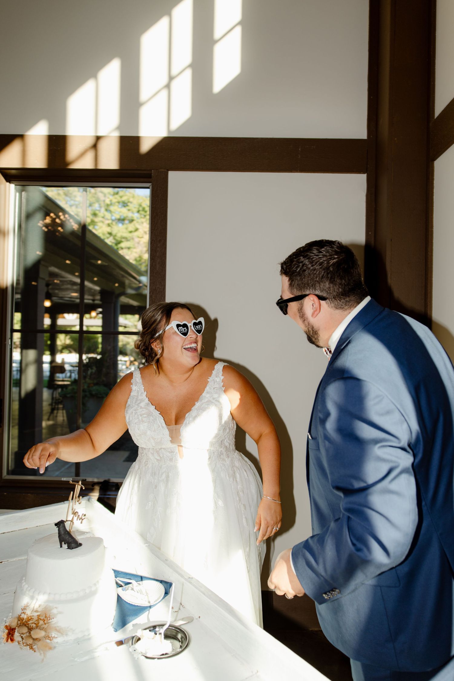 A couple laughs while cutting their wedding cake in a sunlit room.