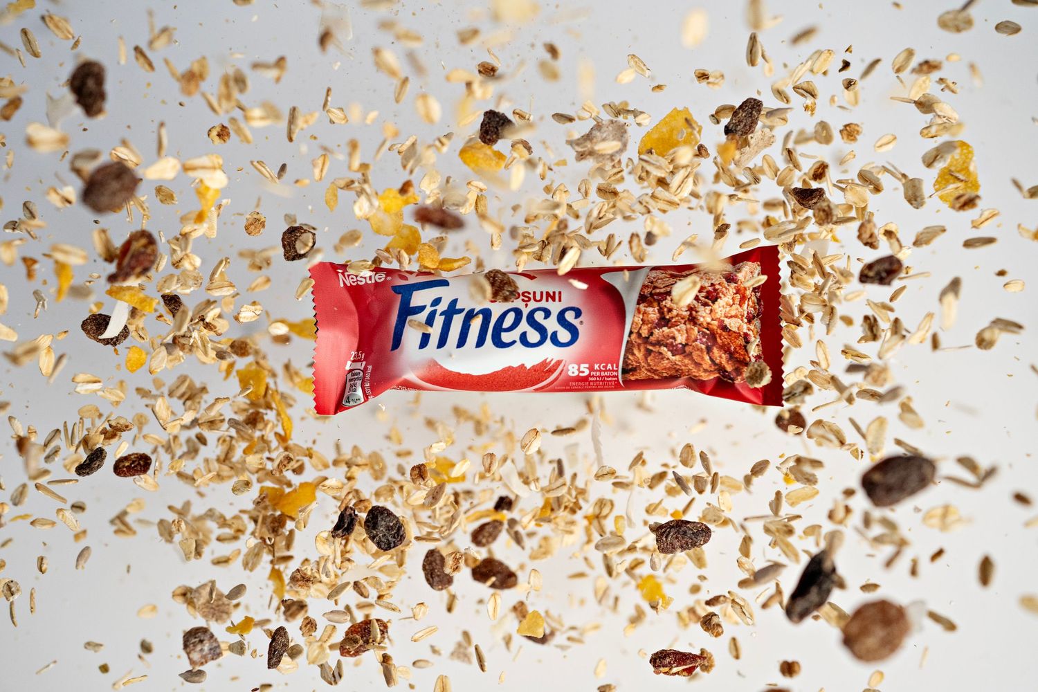 A Nestle Fitness cereal bar surrounded by scattered chocolate pieces and oat flakes on a white background.