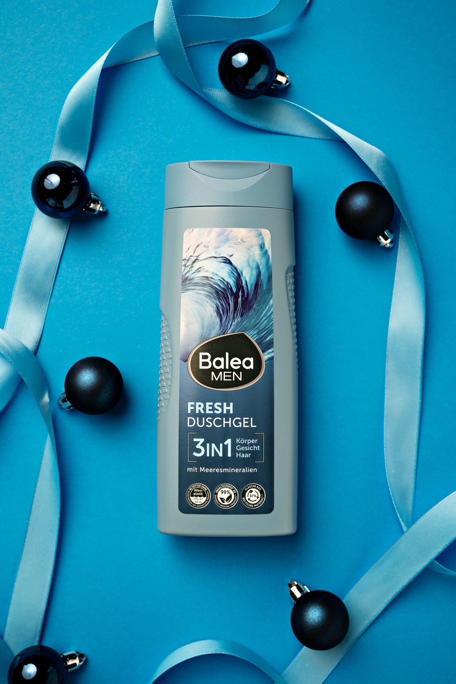 Balea men's grooming products arranged with black Christmas ornaments and blue ribbon on turquoise background.