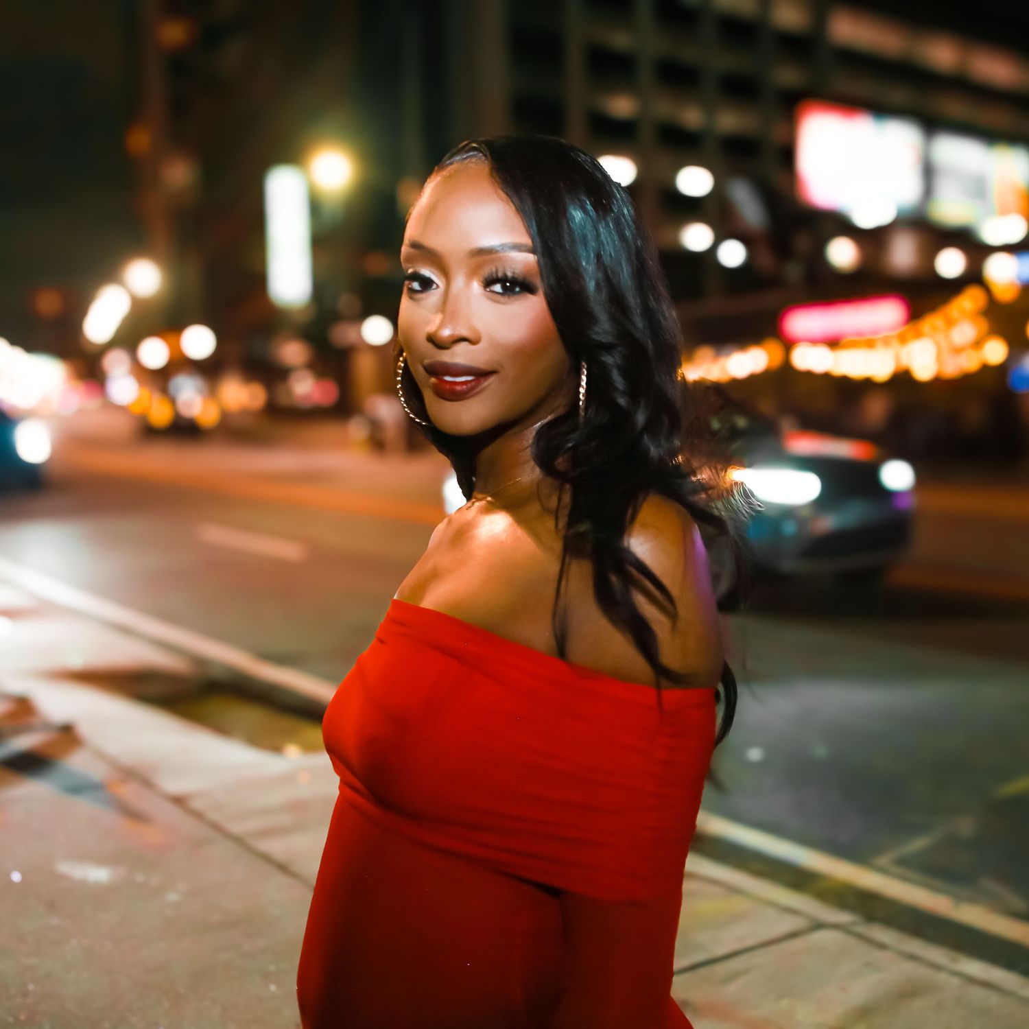 A woman in a red off-shoulder dress poses on a city street at night with bokeh lights.