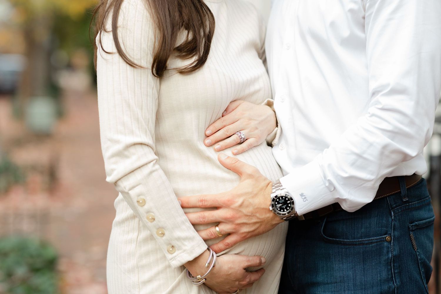 Couple tenderly cradling pregnant belly outdoors in fall, woman in cream sweater dress, man in white shirt.