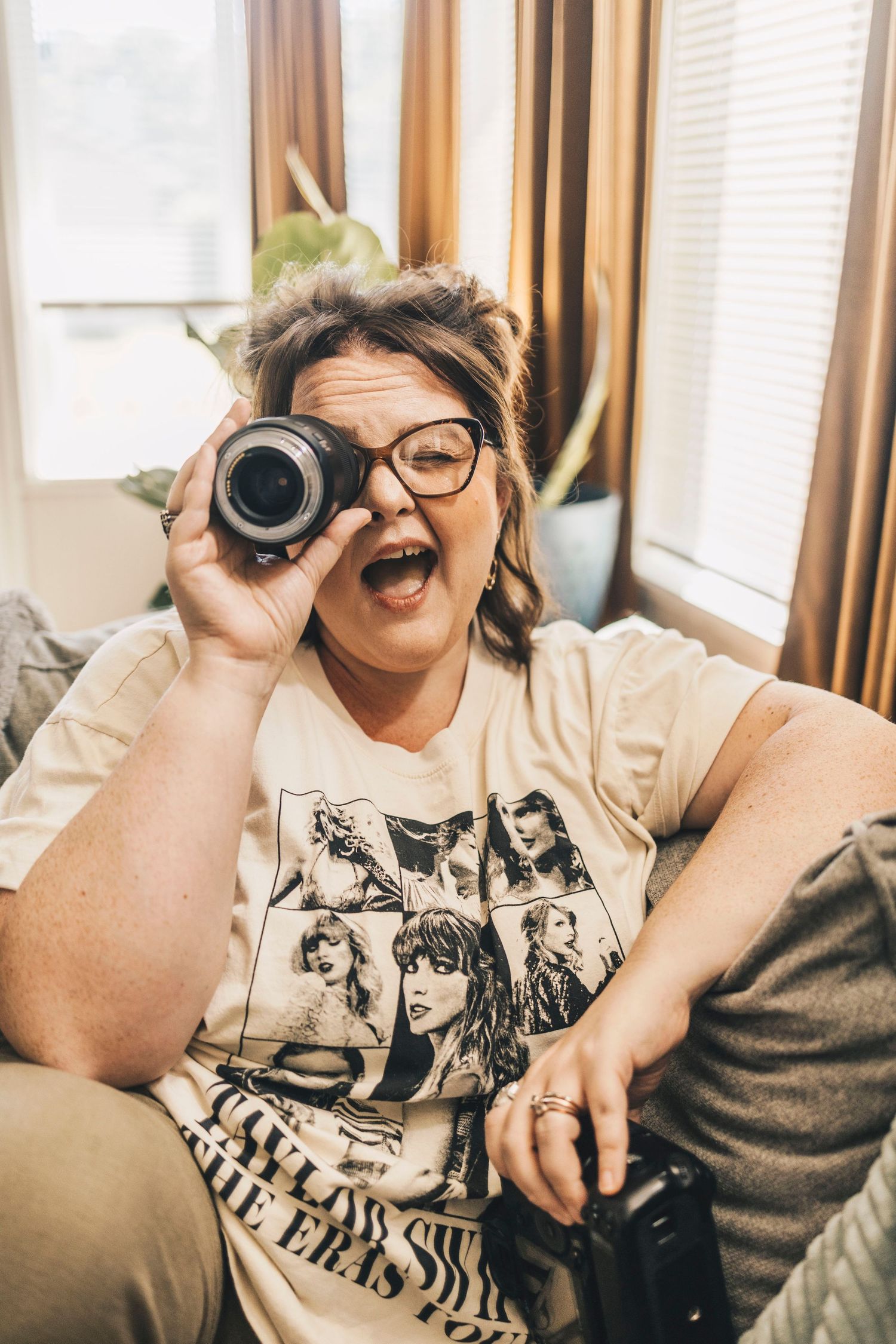 Excited woman in Taylor Swift Eras Tour shirt looking through camera lens on couch.