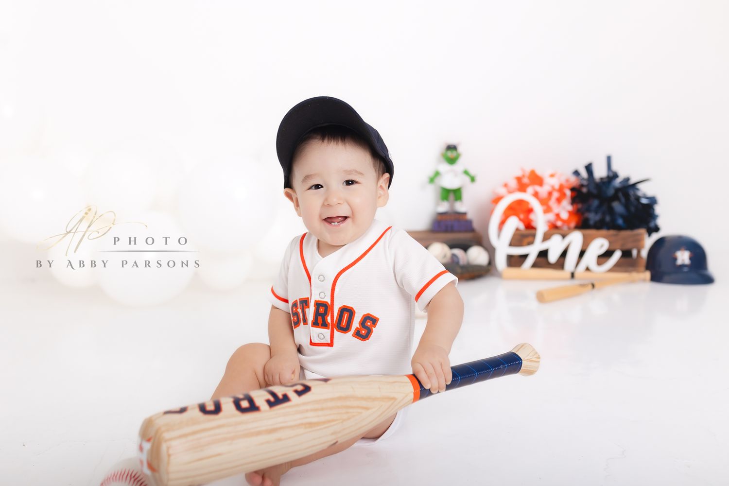 Baby wearing Giants baseball jersey smiles while holding wooden baseball bat with sports equipment in background.
