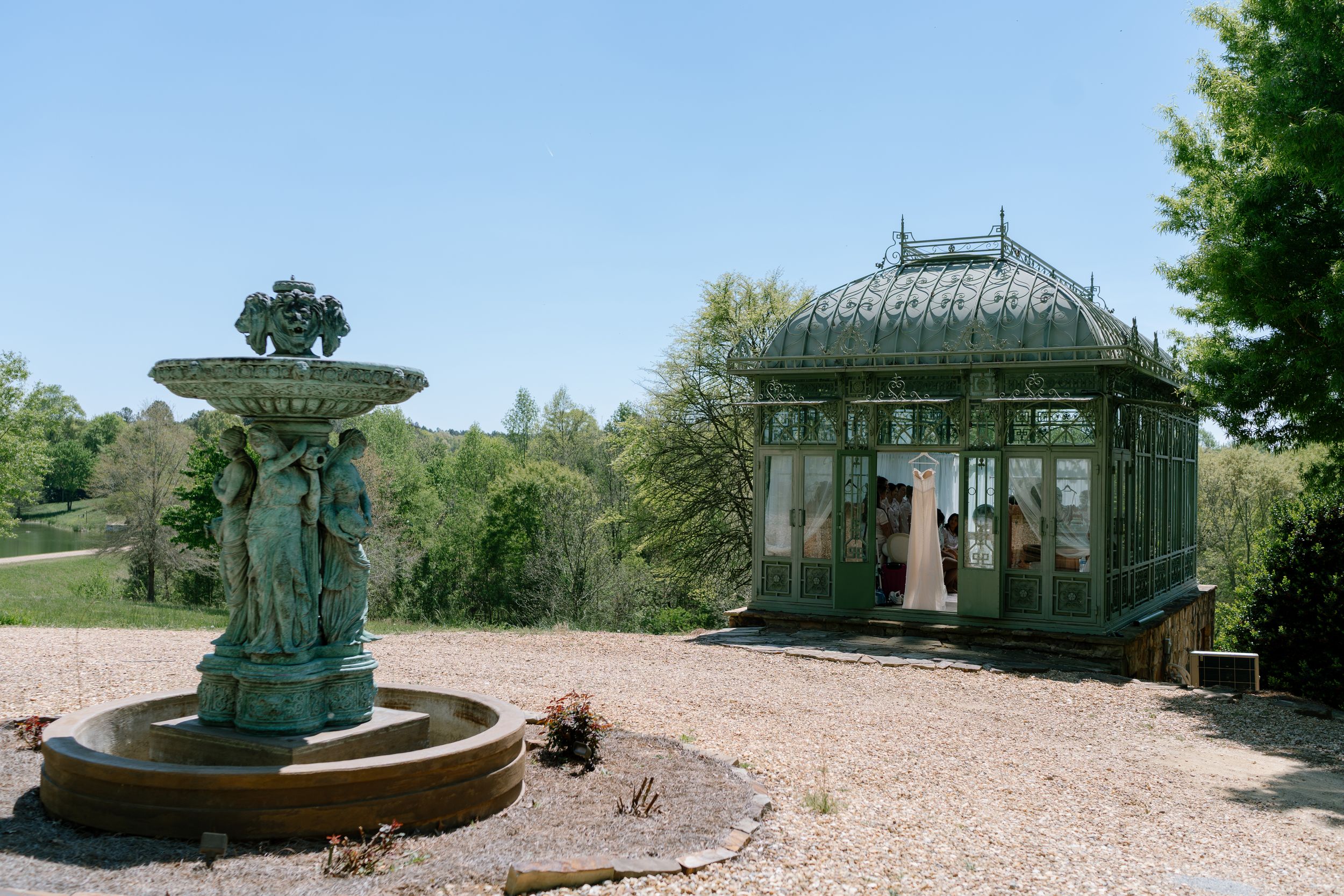 Ornate green bronze fountain with figures beside a Victorian glass gazebo in a landscaped garden setting.