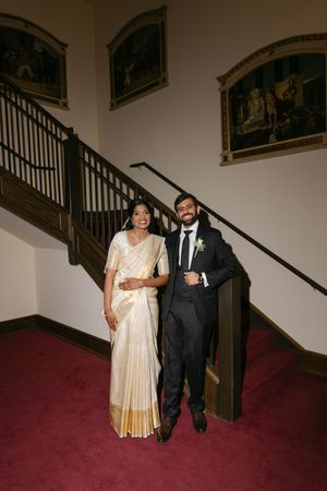 Couple in traditional Kerala wedding attire posing on red carpet staircase, bride in white gold saree, groom in dark suit.