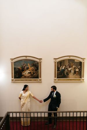 Bride in white saree and groom in black suit hold hands on staircase beside ornate framed paintings.