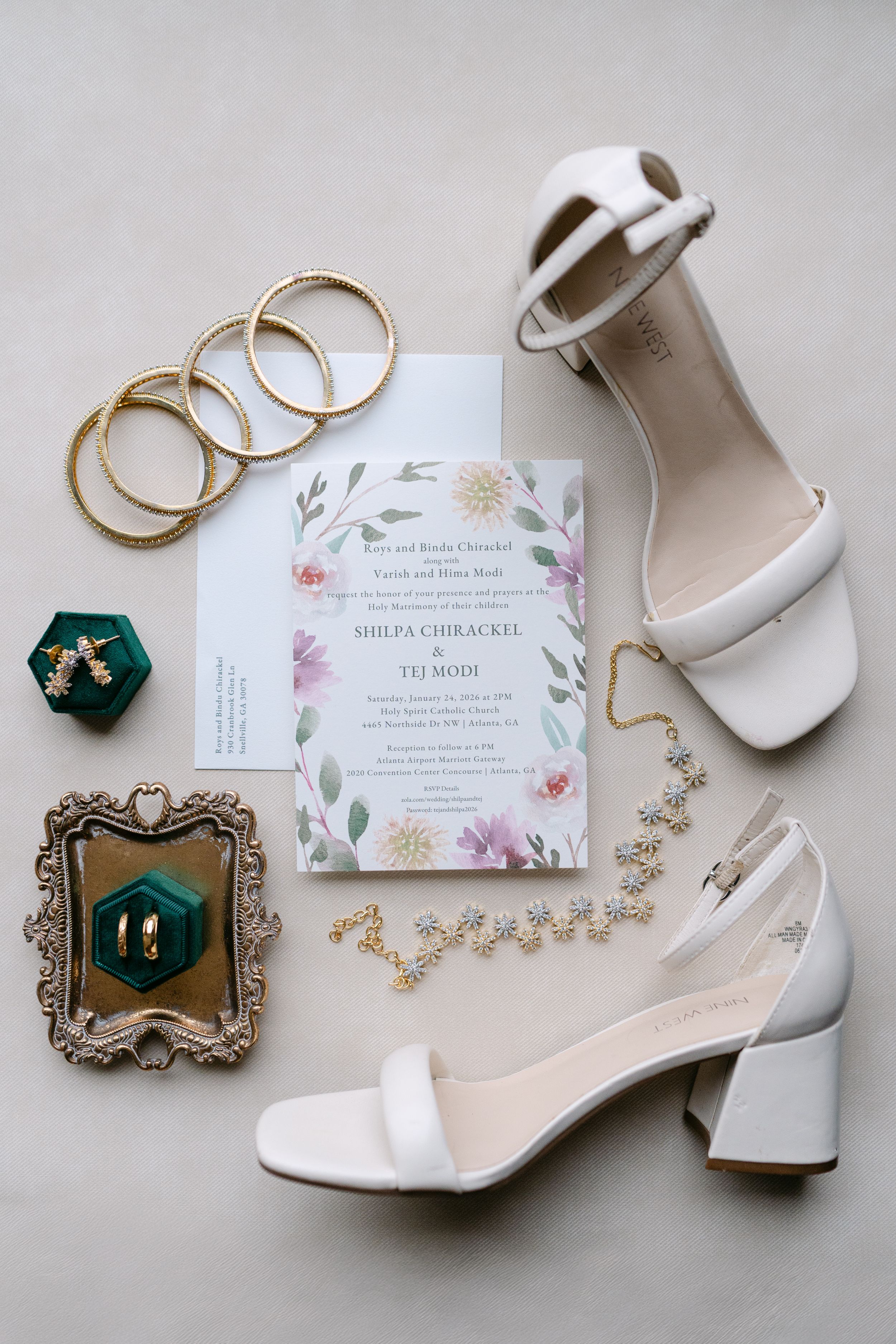 Elegant wedding flat lay featuring white block heels, gold bangles, floral invitation, green velvet ring box, and delicate gold bracelet.