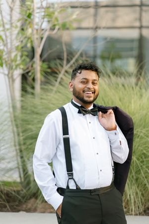 Smiling man in white tuxedo shirt, bow tie, suspenders, holding jacket over shoulder outdoors.