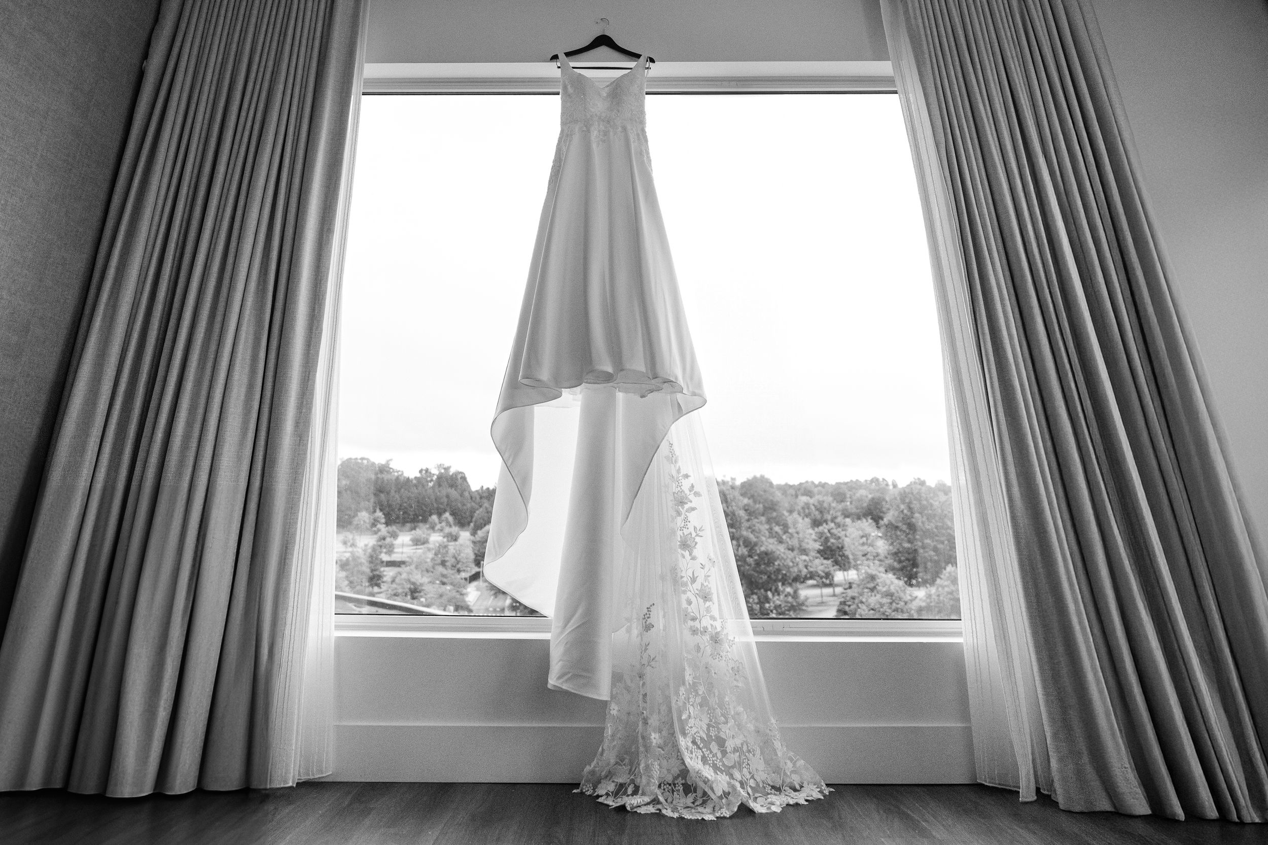A black and white photo of a white wedding dress hanging in front of a window with curtains framing the view.