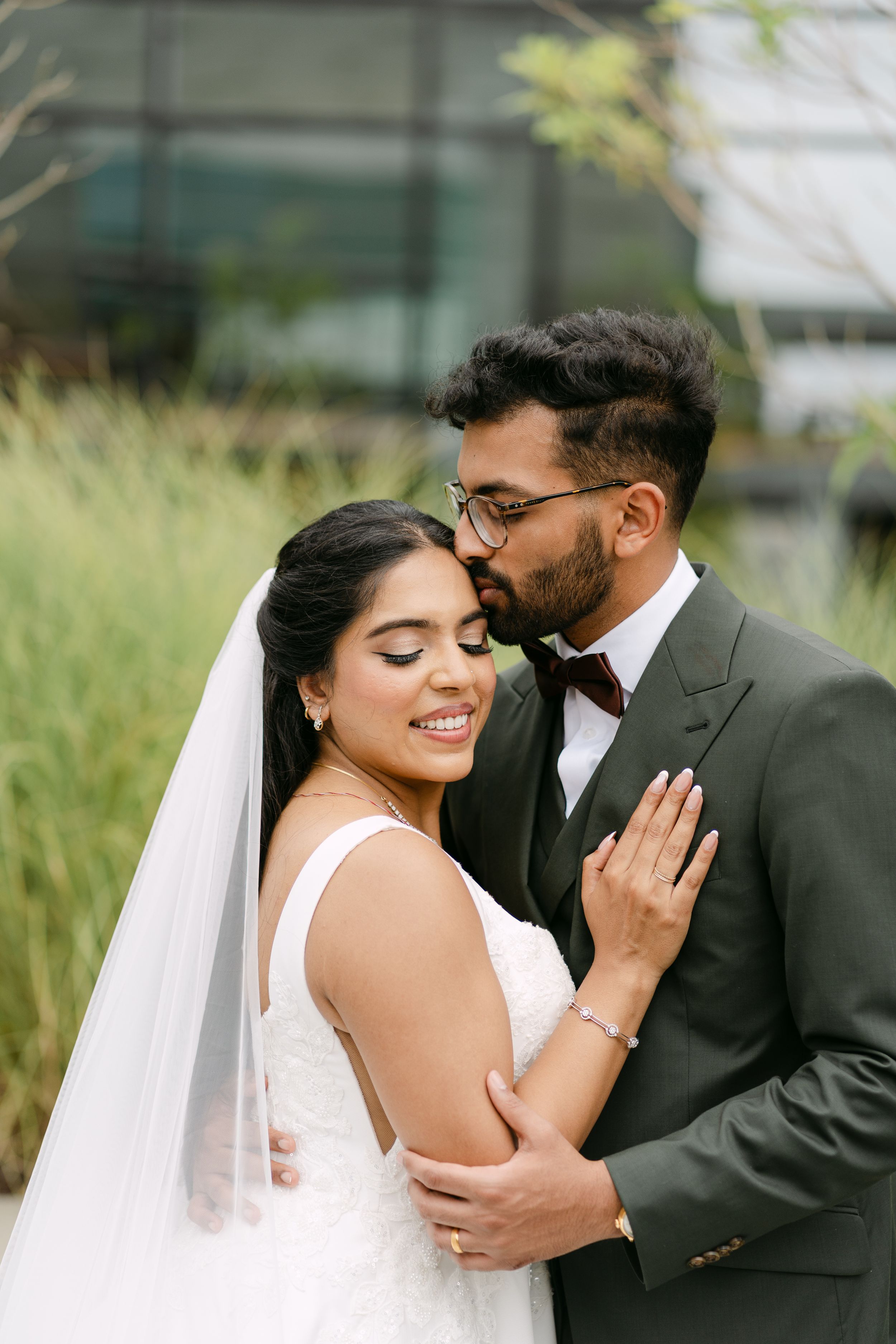 A romantic wedding couple shares intimate moments in a garden setting with tall grass and modern architecture.