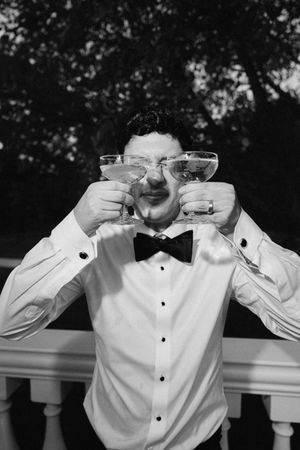 Man in tuxedo shirt and bow tie humorously holds two cocktail glasses up to his face outdoors.