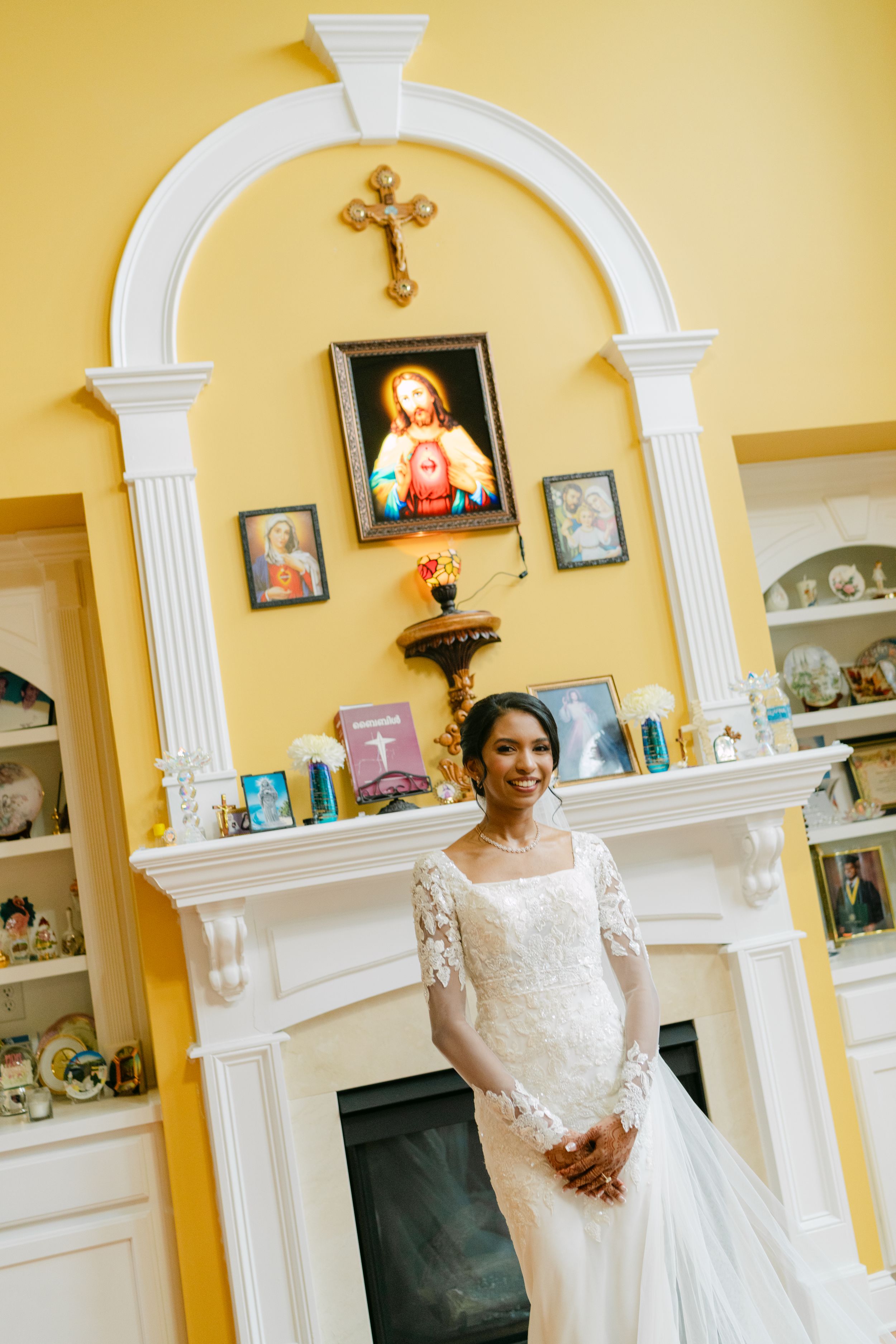 Smiling bride in lace wedding gown stands before white fireplace with religious icons on yellow wall. Syro Malabar Wedding in Georgia.