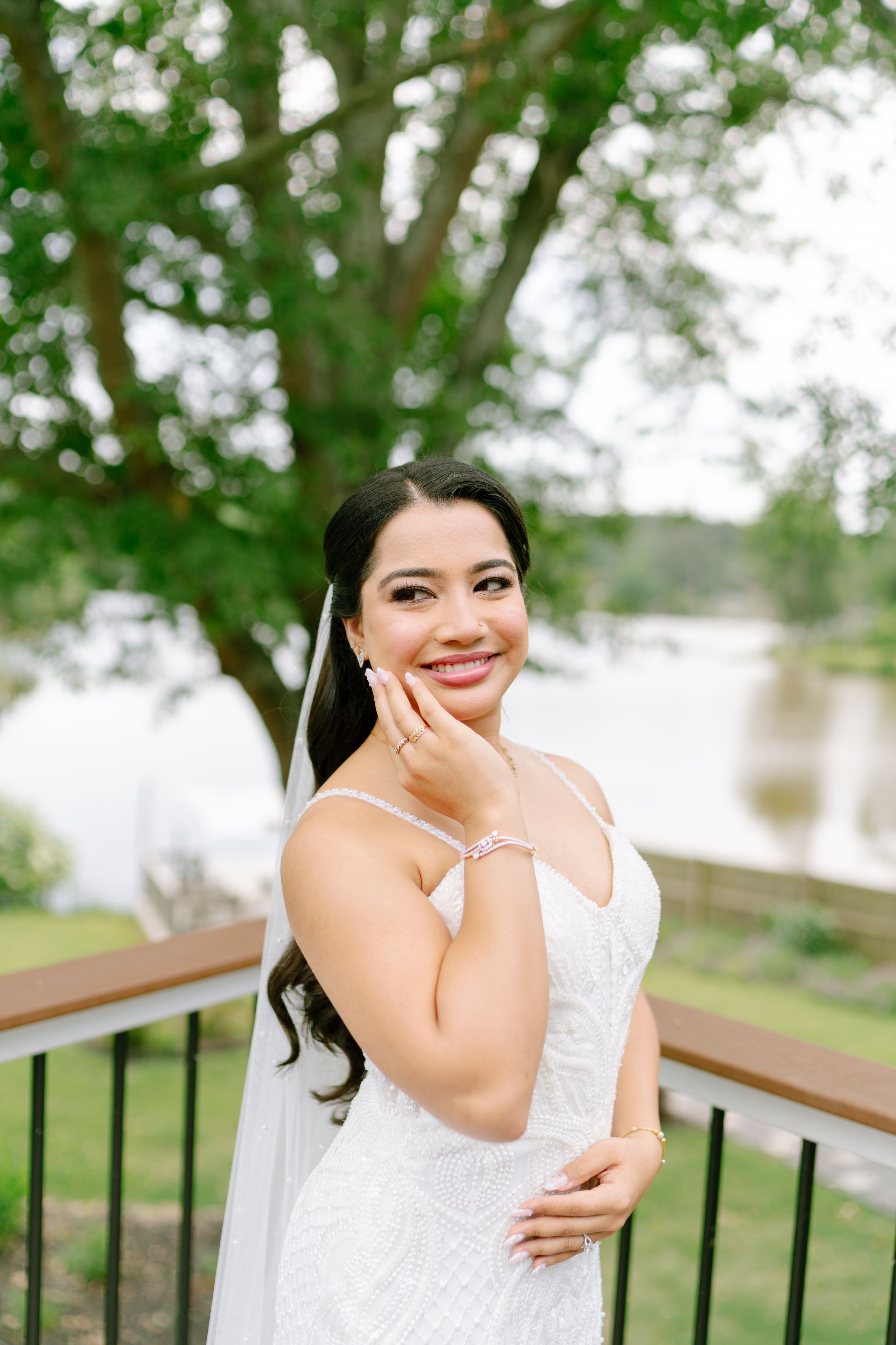 A Bride in a white sleeveless dress poses outdoors on a wooden bench with trees and water in the background. Syro Malabar Catholic Wedding.