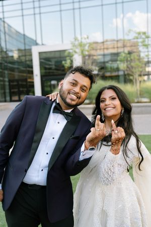 Bride and groom in wedding attire posing outdoors, showing off rings with big smiles near a glass building. Syro Malabar Wedding in Georgia.