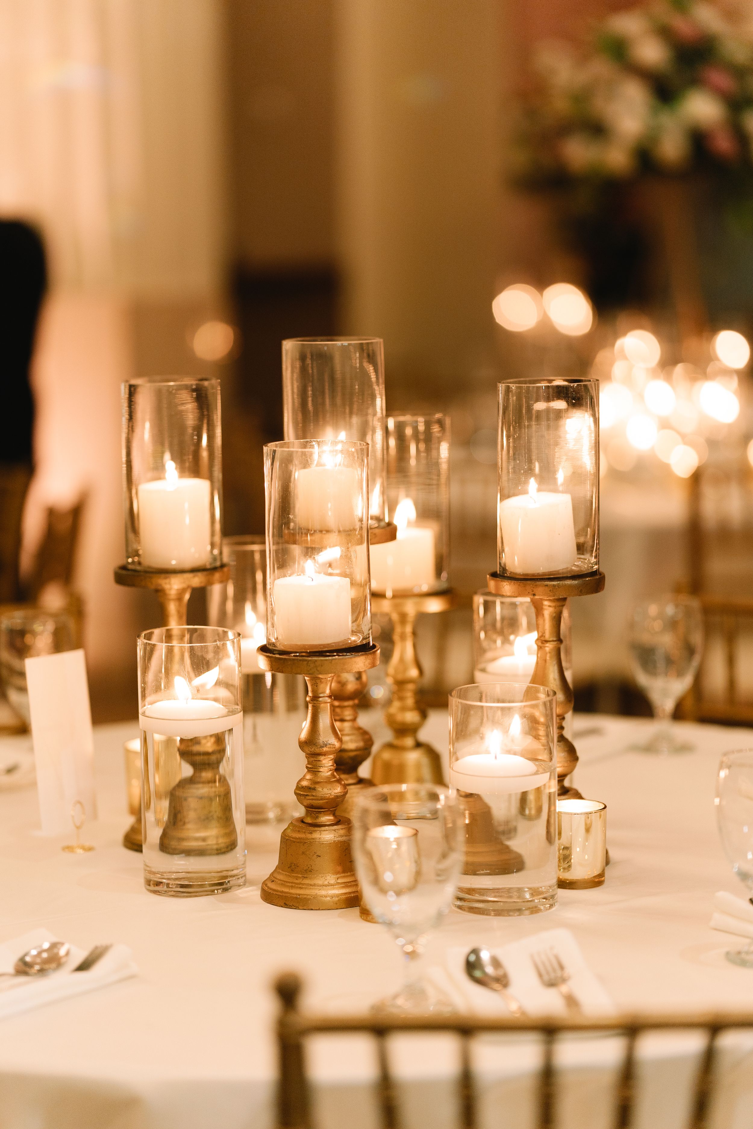 Elegant wedding reception table with gold candleholders, glass hurricanes, and white pillar candles creating warm romantic ambiance.