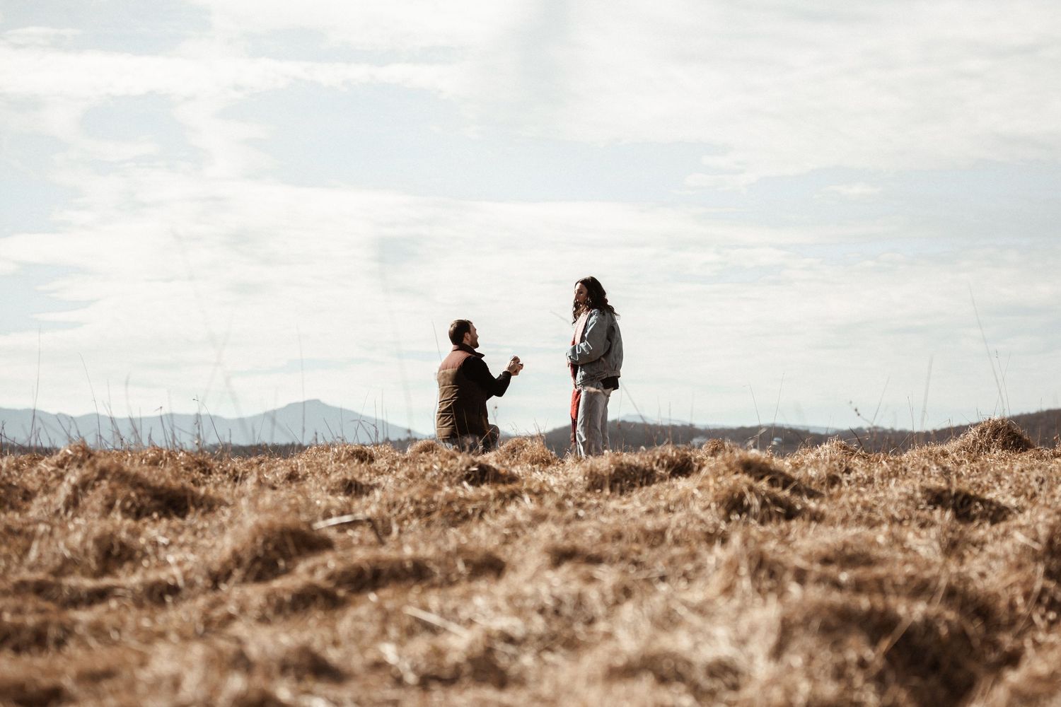 Two silhouettes stand together on a hilltop against a cloudy sky and mountain backdrop.