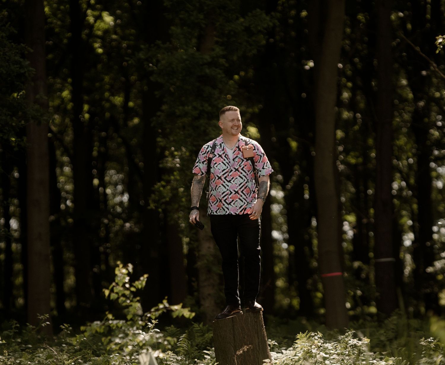 A person in a geometric patterned shirt stands on a log in a moody forest setting.