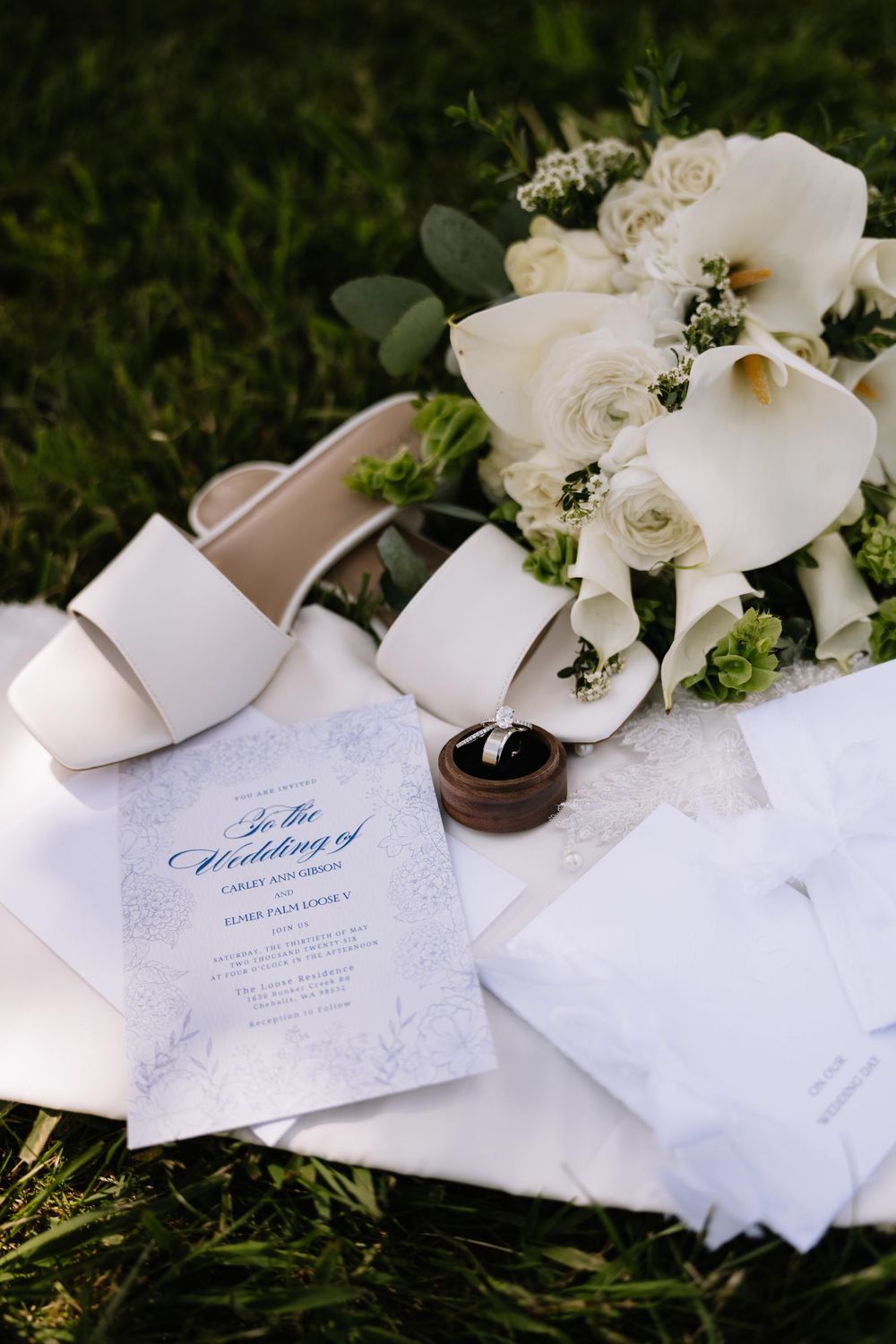 Elegant wedding flat lay featuring white heels, floral bouquet, ring box, and wedding program on green grass.