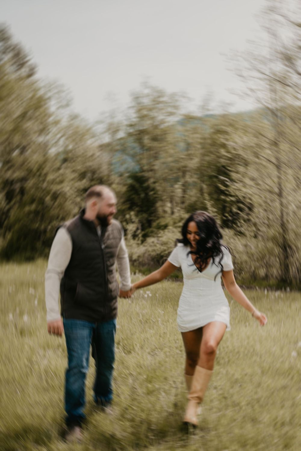 Couple holding hands walking through a lush green meadow, woman in white dress and boots, man in vest and jeans.