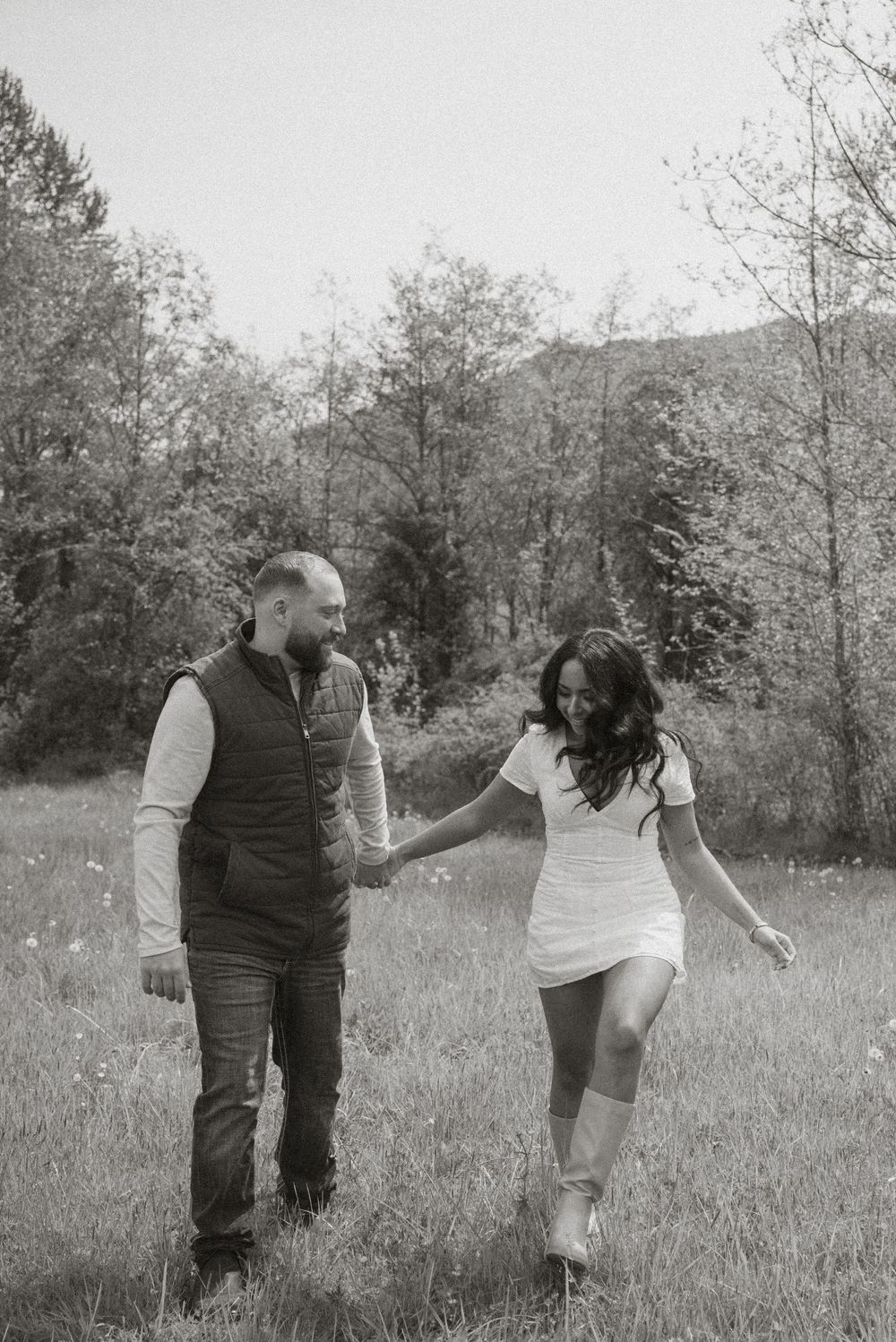 Black and white photo of a couple holding hands and walking through a grassy meadow with trees and mountains in the background.