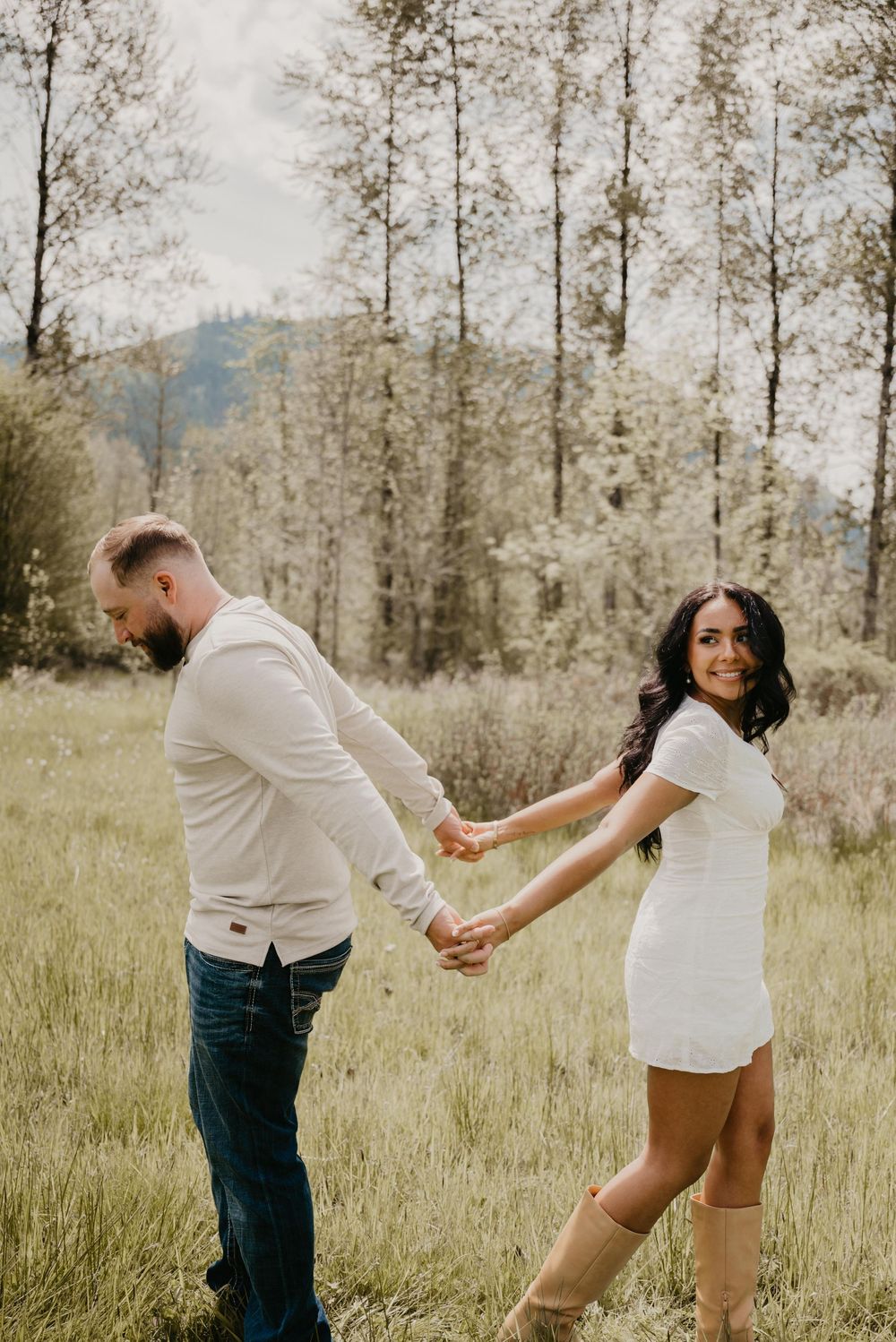 Couple holding hands in a green meadow, woman in white mini dress and boots smiling back at camera, trees in background.