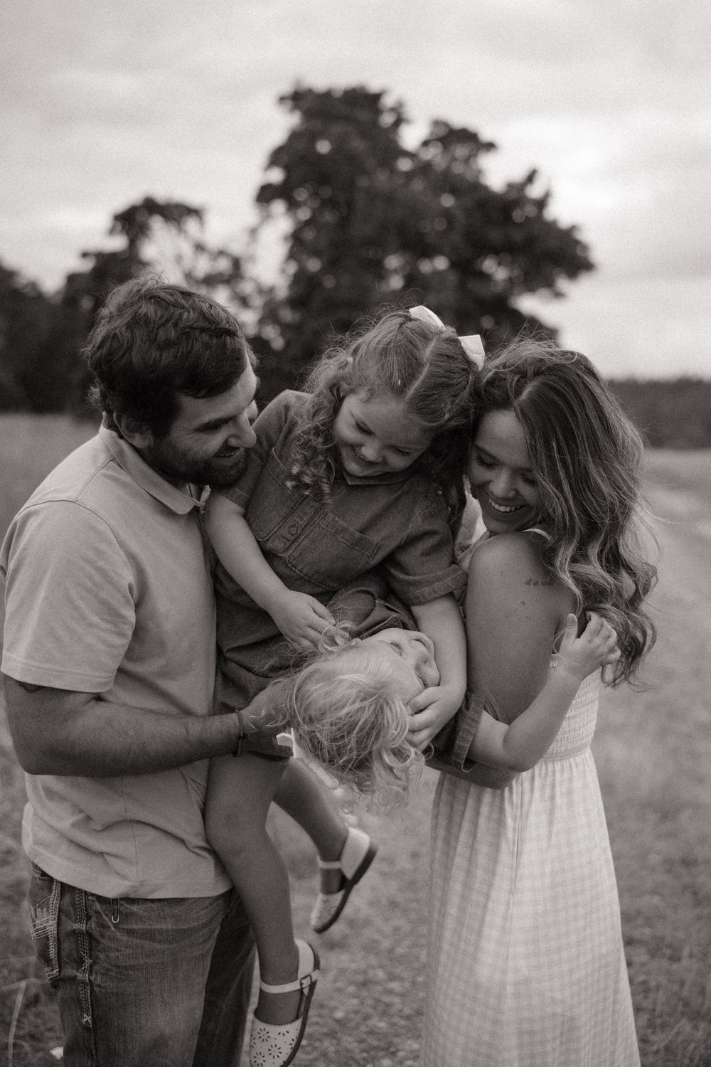 Black and white family photo of parents laughing with two young daughters in an open field with trees in background.
