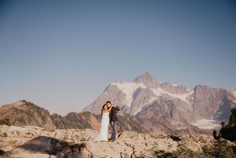 A couple stands on a rocky mountain peak with snow-capped Mount Baker rising majestically in the background.