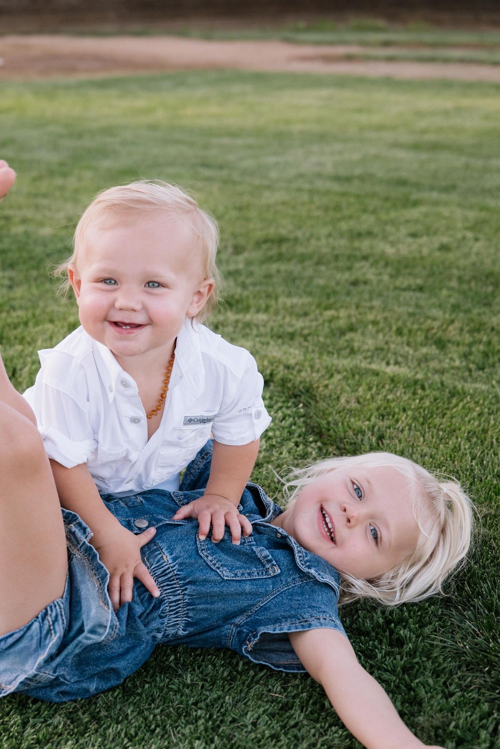 A small child in a white shirt and denim overalls rolls playfully on green grass during a sunny day.