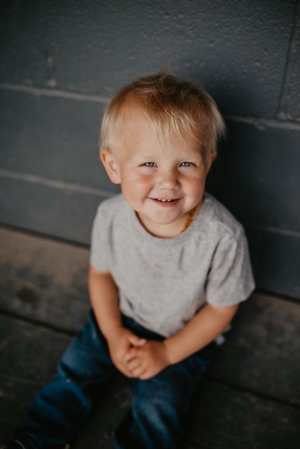 Smiling toddler boy with blonde hair wearing a gray t-shirt and jeans, sitting against a gray wall.
