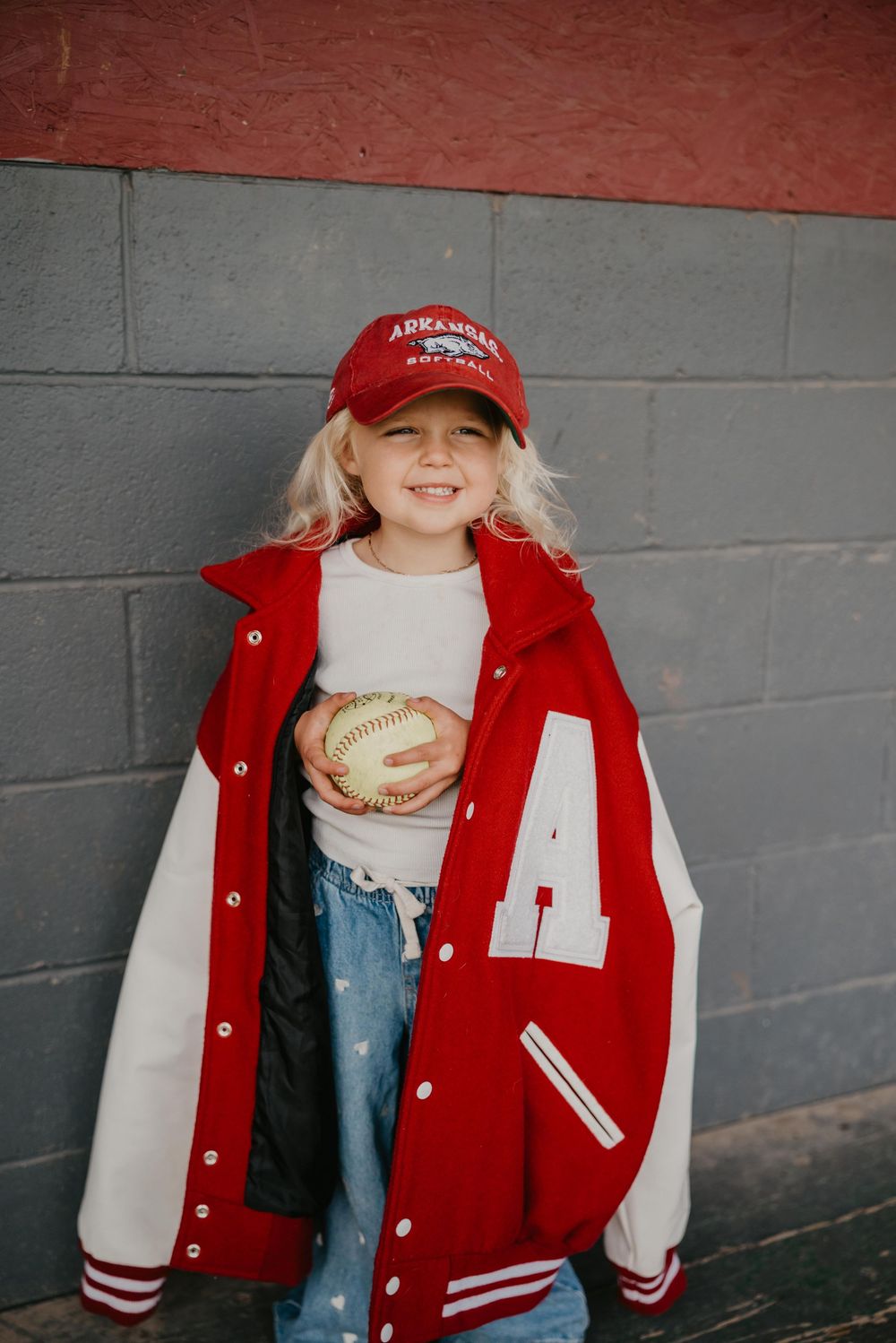 Young girl smiling in red Arkansas varsity jacket and cap, holding a softball against a gray brick wall.