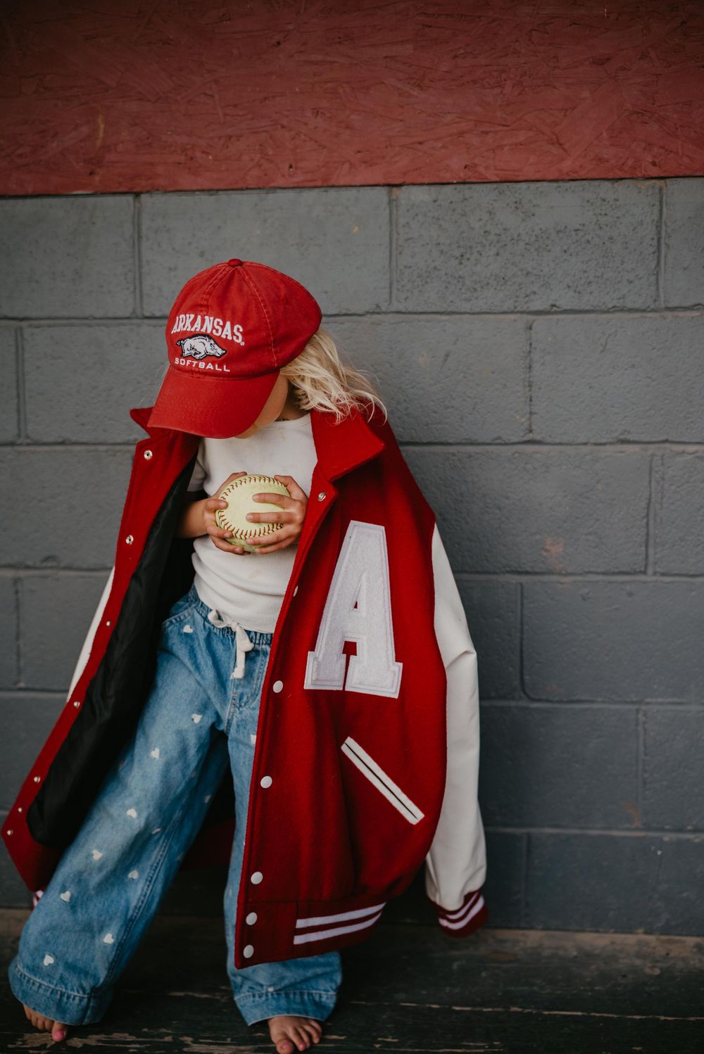 Child in oversized red Arkansas Football varsity jacket and matching cap holds a baseball against a gray brick wall.