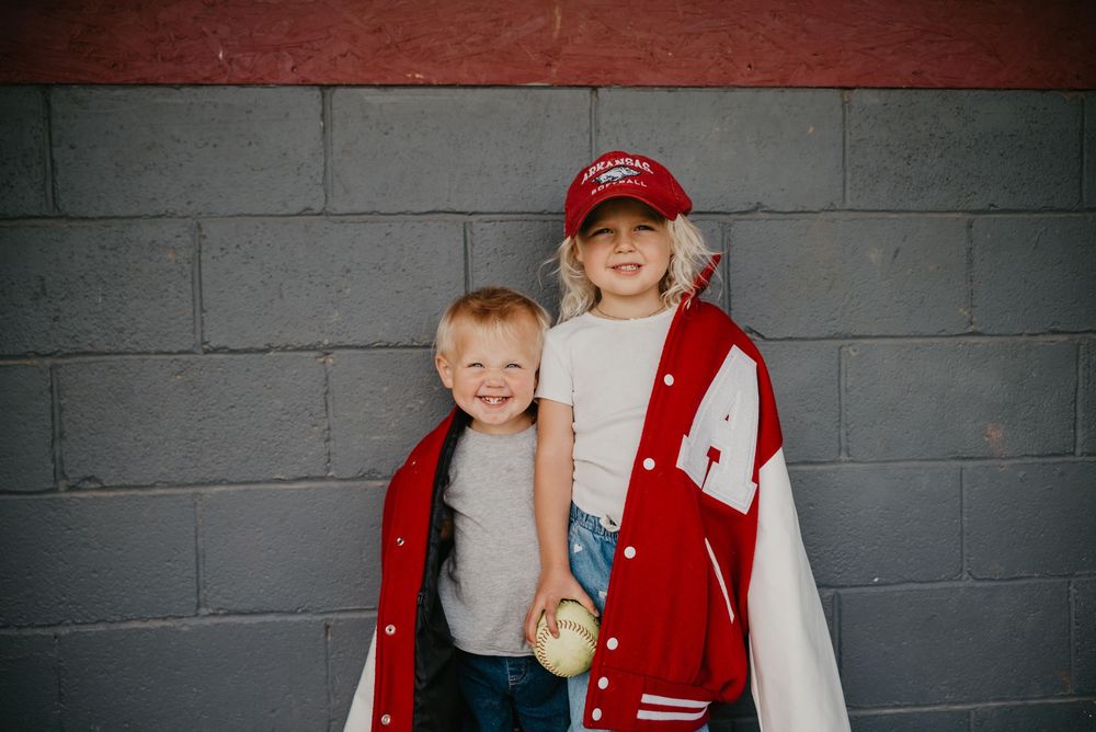 Two young children smiling in red varsity jackets and a red cap posed against a gray brick wall.