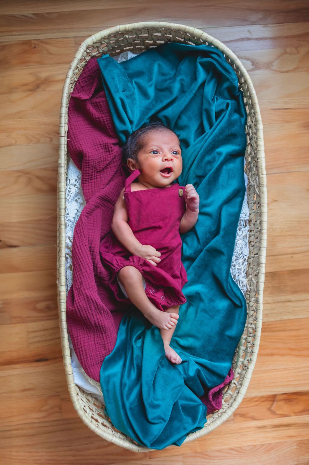 Birds eye view of a newborn baby in a moses basket.  He is wearing a burgundy romper.  There is a green blanket and a burgundy blanket.