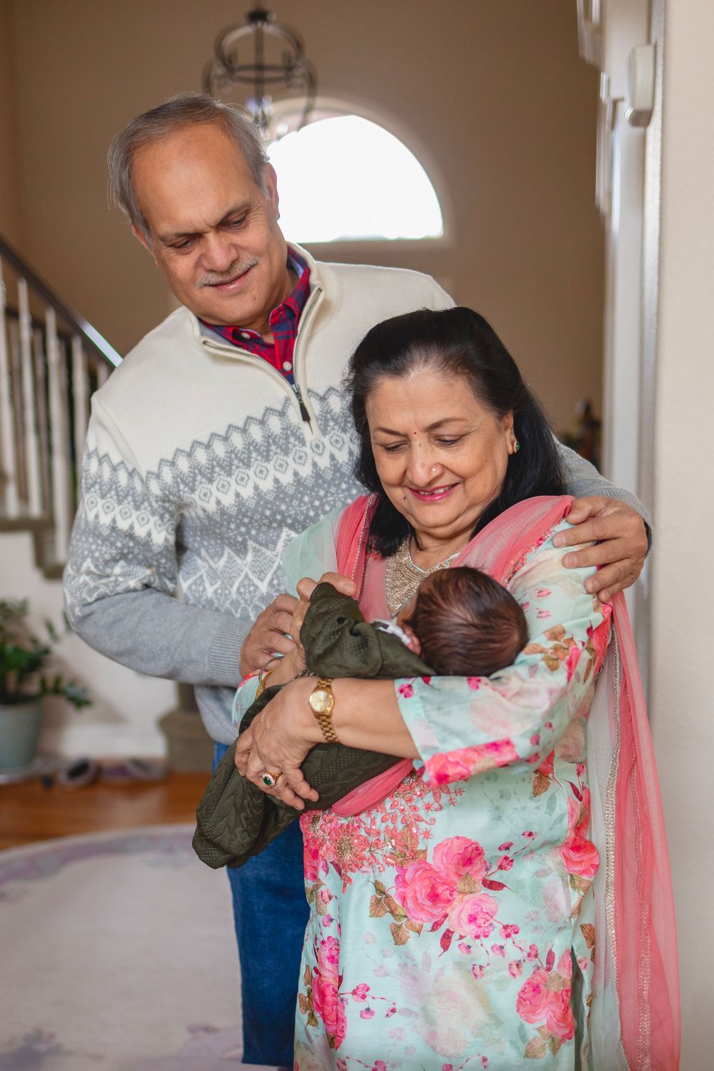 Grandmother holding newborn baby, Grandfather is stood behind her with his arm around her, he is looking down at baby.