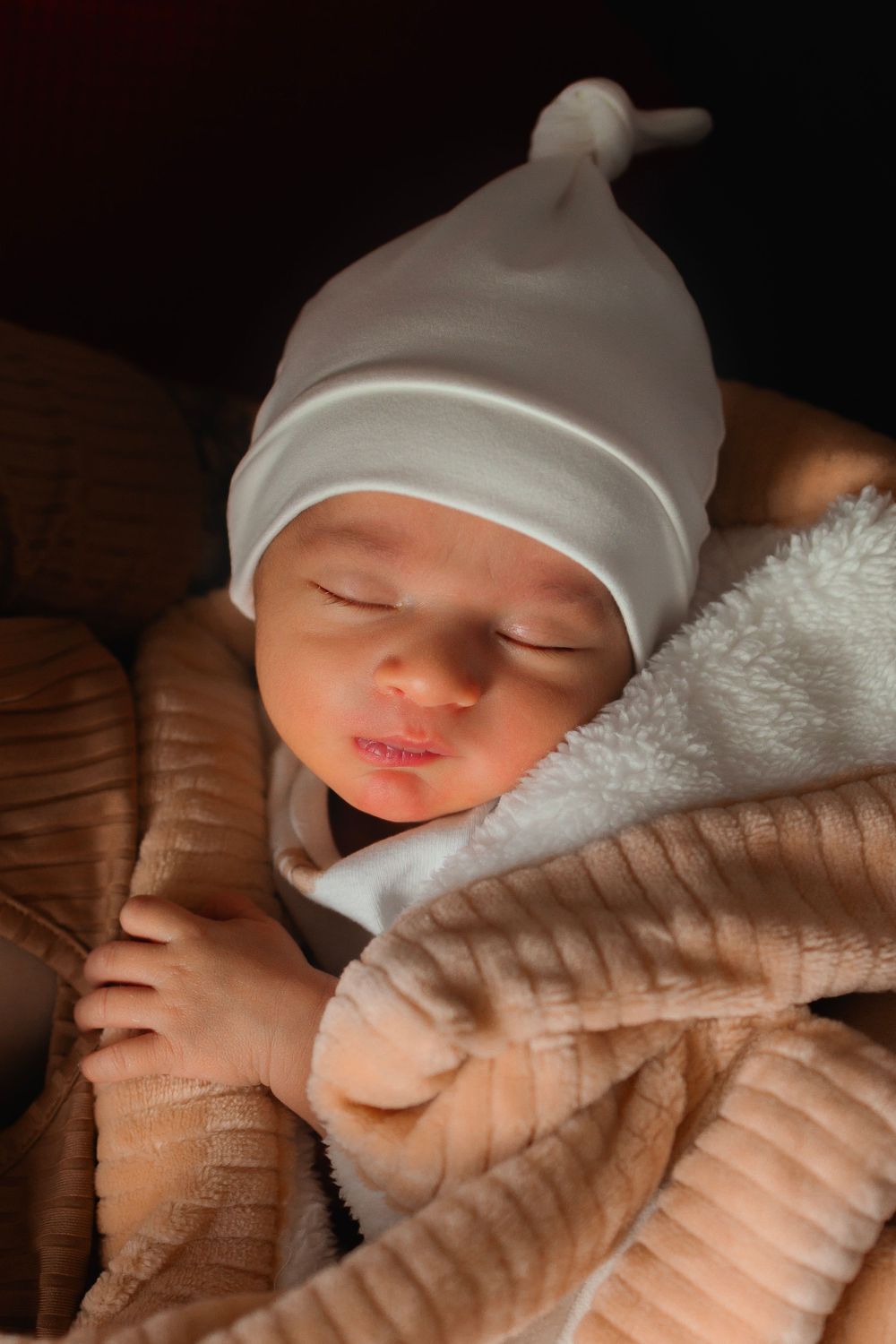 Sleeping newborn baby wearing white knotted cap snuggled in soft brown and white blankets.
