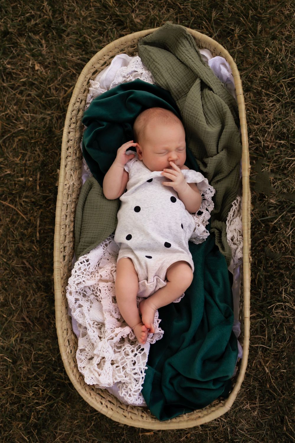 Newborn in white lace dress sleeps peacefully in woven basket with green blanket shot from above.