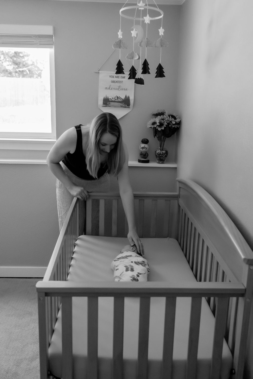Woman leaning over and laying her hand on sleeping newborn inside a gray wooden crib beneath a mobile with star decorations in a nursery.