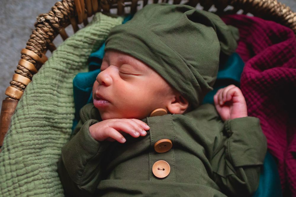 Sleeping newborn wearing cozy olive green outfit sleeps peacefully in woven basket on colorful knit blankets.