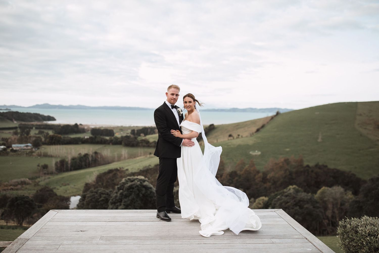 Newlyweds standing on wooden deck overlooking scenic coastal hills and ocean view.