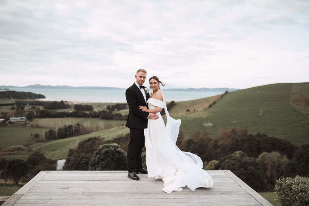 Newlyweds standing on wooden deck overlooking scenic coastal hills and ocean view.