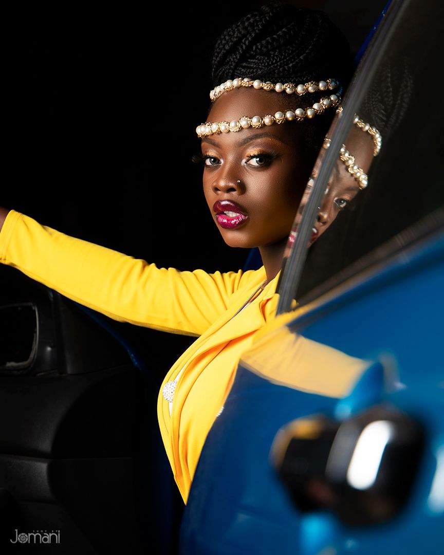 Woman in yellow long-sleeve top with pearl headpiece poses beside a blue car in dramatic lighting.