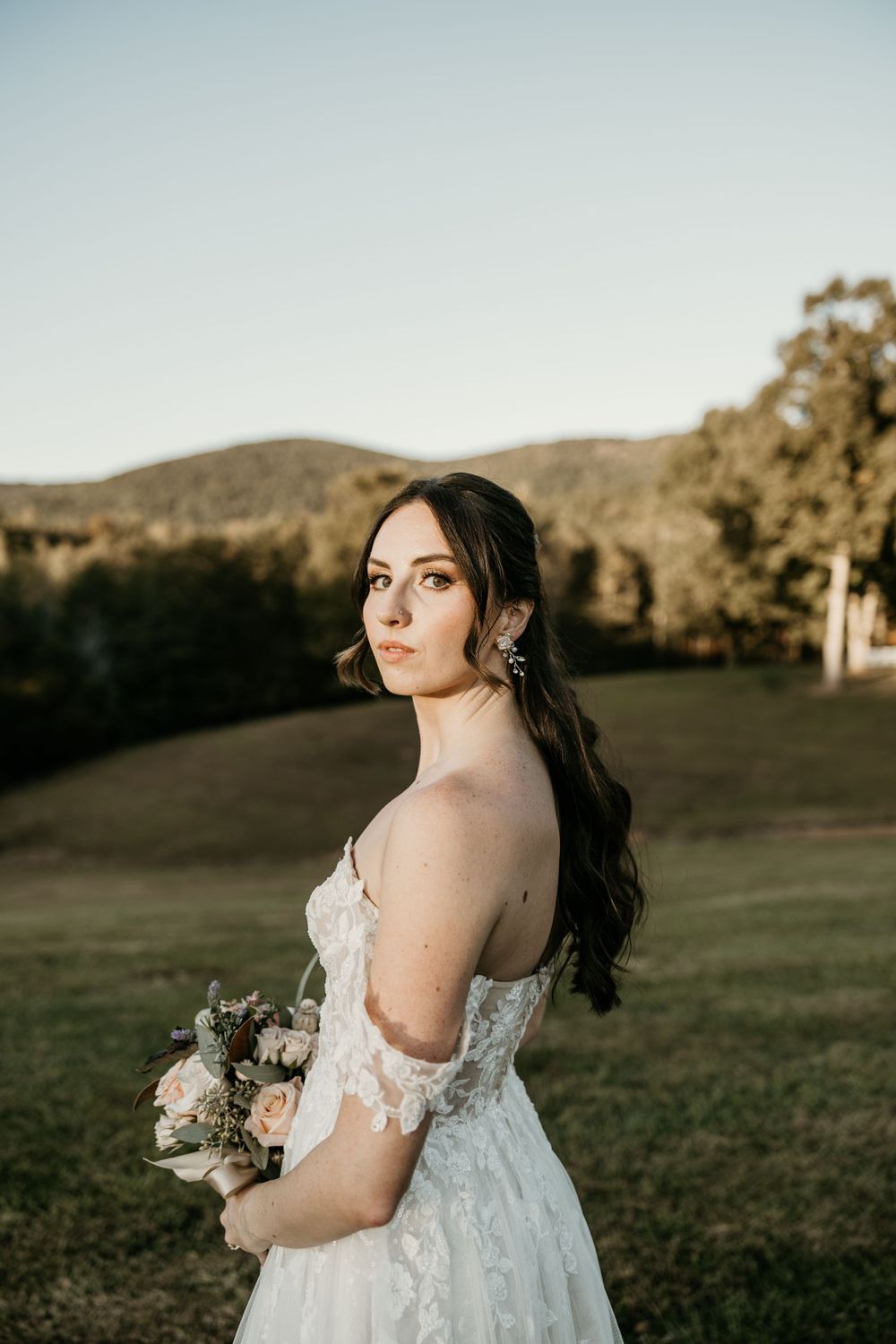 Off-shoulder lace wedding dress with bouquet photographed against rolling hills at golden hour.