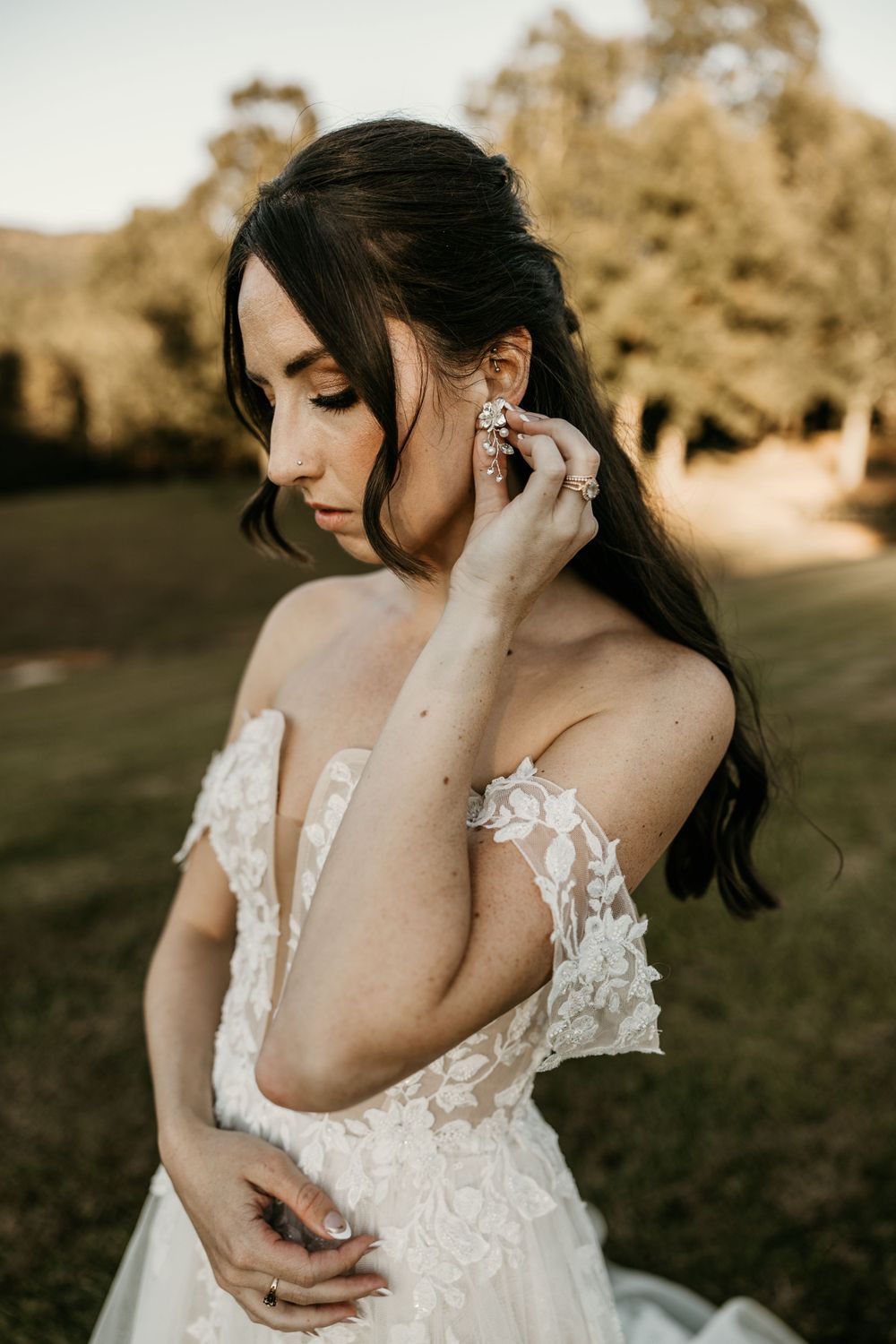A bride in lace gown adjusts her earring in golden sunset light.