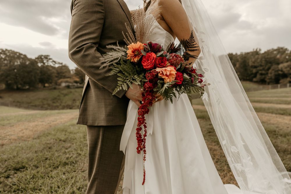 Wedding couple holds vibrant burgundy and coral floral bouquet against cloudy countryside backdrop in outdoor ceremony.