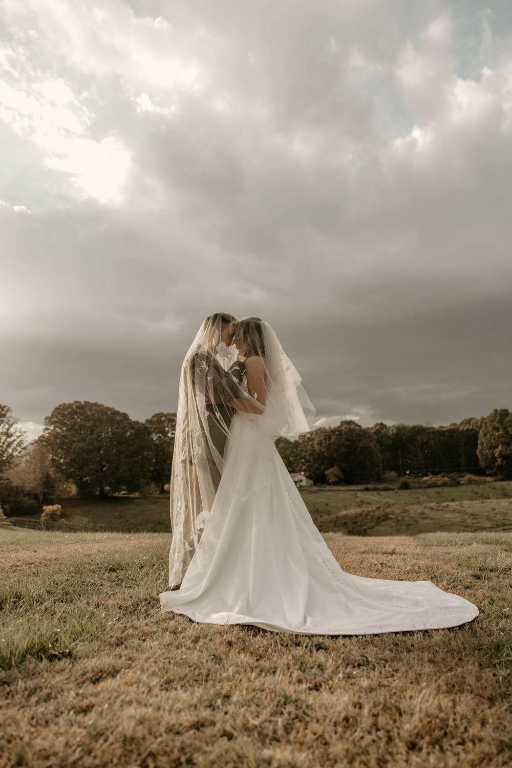 A bride in a flowing white wedding gown with a long veil stands in a field under dramatic cloudy skies at sunset.