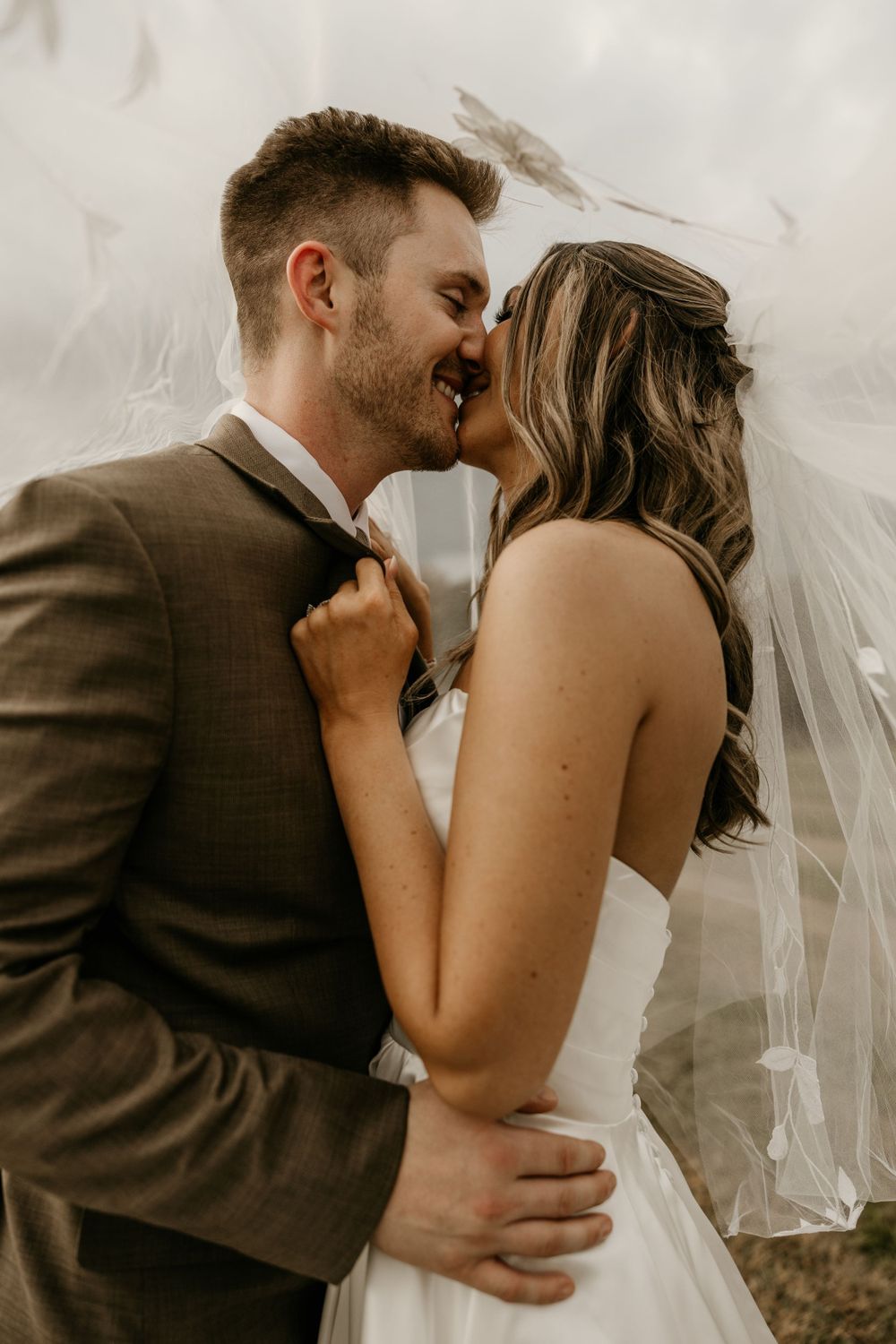 Romantic wedding portrait of couple embracing as veil blows in wind against neutral background.