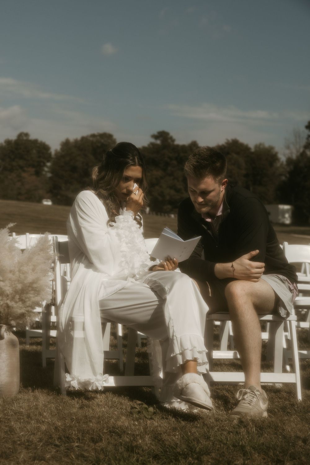 Two people sit together on white chairs outdoors reading from a piece of paper.